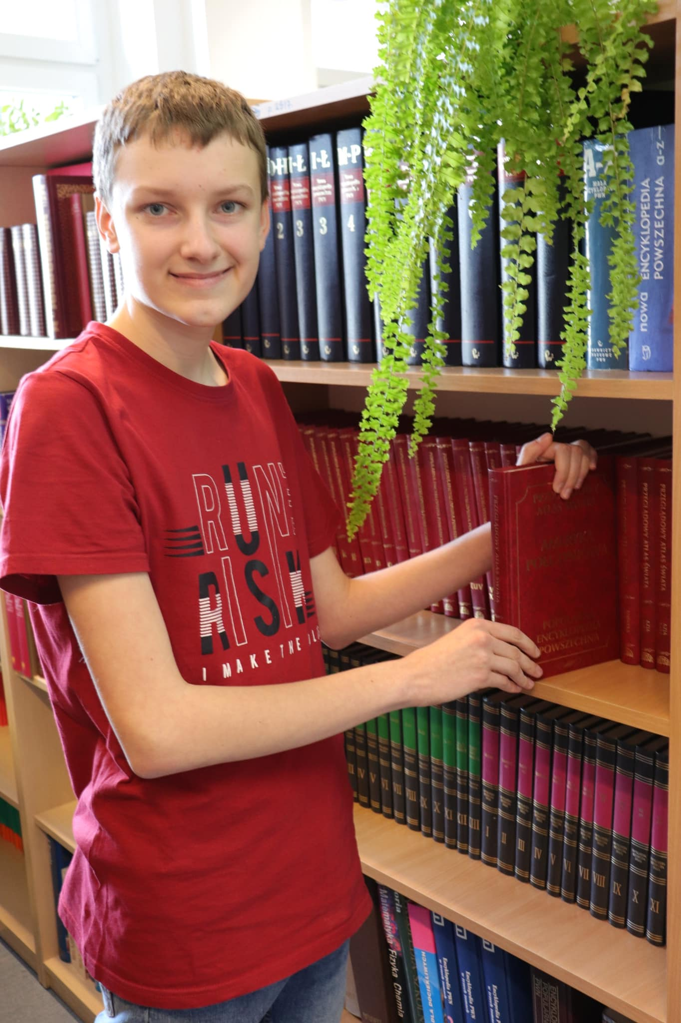 Stanisław Kudzia Zdolny Ślązak ze SP w Pszennie. Stoi na tle półki z książkami i jedną z nich trzyma w ręku. na półce stoi paproć