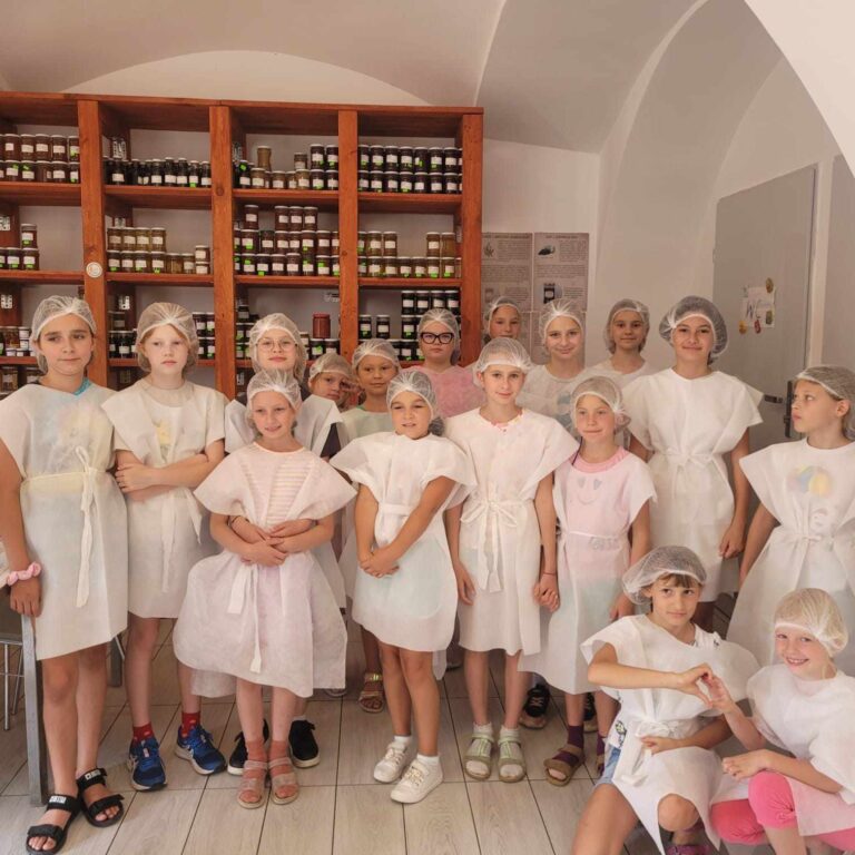 Uczestnicy warsztatów serowarskich stojący w fartuszkach i czepkach na głowę a tle regałów z produktami spożywczymi