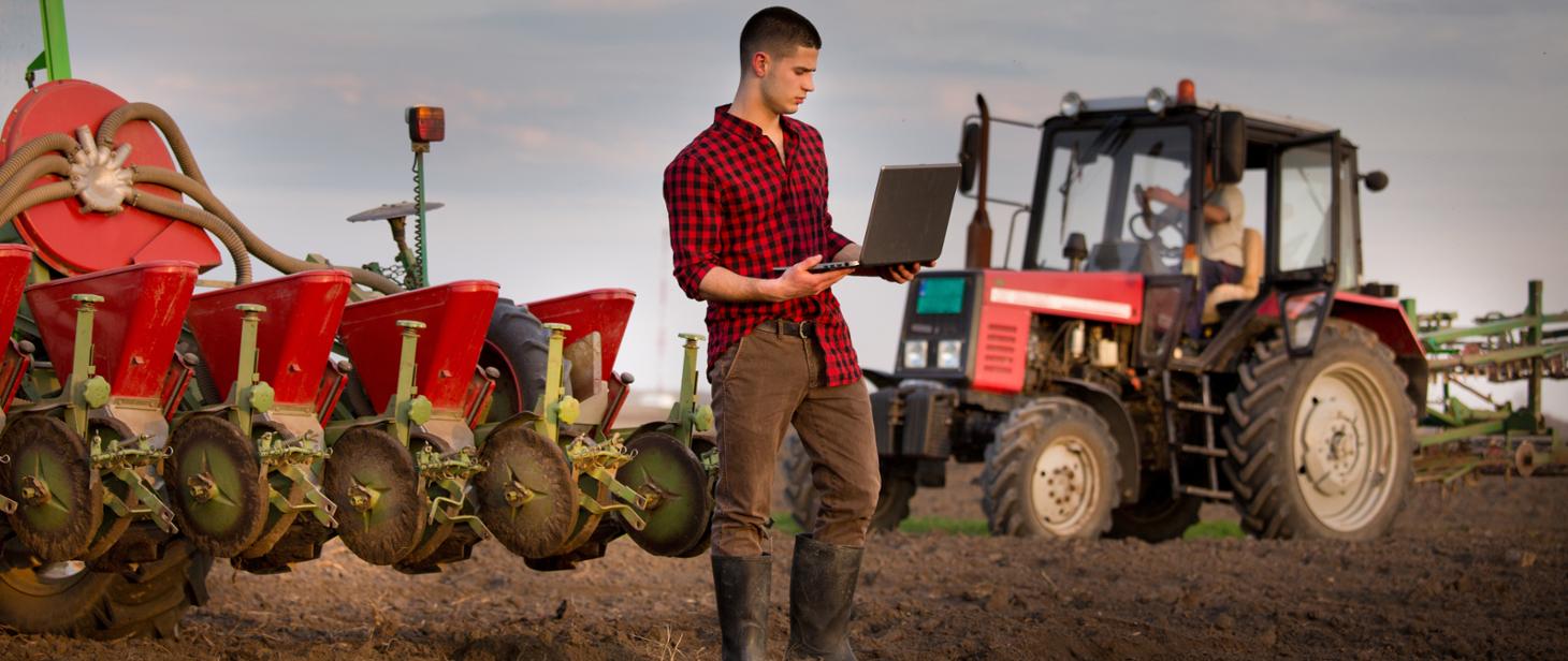 Rolnik trzymający w ręce laptop, w tle maszyny rolnicze