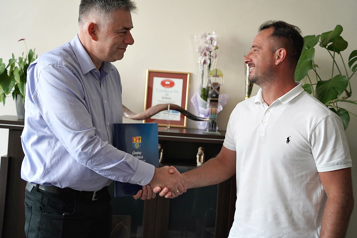 Przekazanie podpisanej umowy na przebudowę drogi gminnej w siedzibie Urzędu. Od lewej wójt gminy Bartłomiej Strózik, od prawej wykonawca Emil Laskowski. W tle komoda na której są w ramkach dyplomy i kwiaty w doniczkach